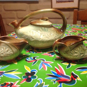 May include: A vintage tea set with a green and brown glaze. The teapot has a unique curved handle and a lid with a brown knob. The creamer and sugar bowl have matching designs.