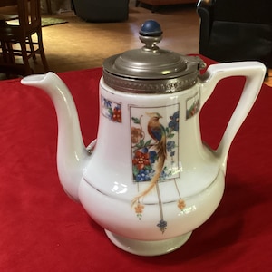 Könnte beinhalten: Weiße Porzellan-Teekanne mit Vogel- und Blumenmuster. Die Teekanne hat einen silbernen Deckel und einen blauen Knopf. Der Griff und der Ausguss sind gebogen. Die Teekanne steht auf einer roten Oberfläche.