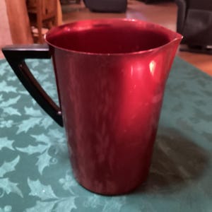 May include: A red pitcher with a black handle. The pitcher is made of a shiny material and has a simple, functional design. It is likely used for serving beverages. The handle is a contrasting black color.
