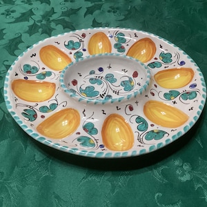 May include: A white ceramic serving platter featuring a central circular bowl surrounded by six oval-shaped indentations. The dish is decorated with hand-painted yellow oval motifs and teal floral patterns with black line accents along the rim.