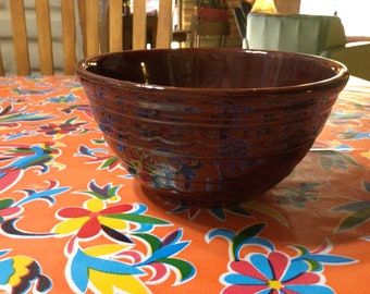 May include: A brown ceramic mixing bowl with a decorative design. The bowl is sitting on a colorful tablecloth with floral patterns.