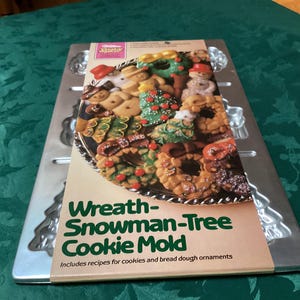 Könnte beinhalten: Eine silberne Wreath-Snowman-Tree Keksform aus Metall mit einem Pappetikett, das ein farbenfrohes Foto von dekorierten Weihnachtsplätzchen zeigt. Der Text lautet Wreath-Snowman-Tree Cookie Mold und enthält Rezepte für Kekse und Brotteigschmuck.
