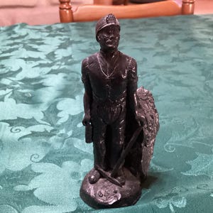 May include: A dark grey metallic figurine of a coal miner wearing a hard hat, a waistcoat, and trousers. The figure stands on a circular base, holding a pickaxe in one hand and a small lantern in the other, with a textured rock formation behind them.