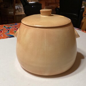 以下が含まれることがあります： ふたと2つの取っ手がついたベージュの陶器製の鍋。鍋は滑らかで丸みを帯びた形をしています。