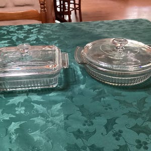 May include: Two clear glass casserole dishes with lids. One is rectangular with handles, the other is round with handles. Both have clear glass lids with small knobs. The dishes are on a green tablecloth.