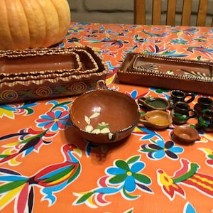 May include: A set of terracotta serving dishes in various sizes, including a large bowl, rectangular platters, and small cups. The dishes are decorated with brown, white, and green paint. The dishes are arranged on a colorful tablecloth with a floral pattern.