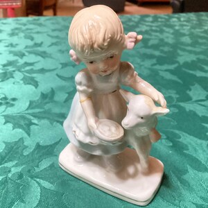 May include: A porcelain figurine of a young girl in a white dress, holding a small lamb. The girl has blonde hair and is standing on a rectangular base. The figurine is painted in soft, pastel colors.