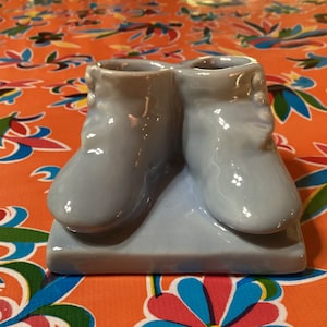 Könnte beinhalten: Ein Paar hellblaue Baby-Schuhe aus Keramik, die an den Fersen miteinander verbunden sind, mit einer Basis zum Aufbewahren von Kleinigkeiten. Die Schuhe haben eine glänzende Oberfläche.