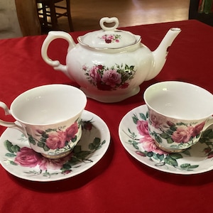 May include: A white porcelain tea set featuring a teapot, two teacups, and saucers. The set is decorated with pink roses and green leaves. The teapot has a heart-shaped handle on the lid. The set is on a red tablecloth.
