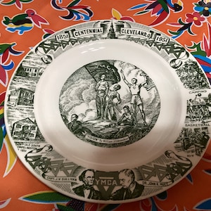 May include: A white ceramic plate with a green and white illustration. The plate features images of buildings, people, and text that reads "1851 Centennial Cleveland, O 1951" and "YMCA". The plate is on a colorful patterned surface.