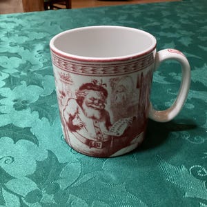 May include: White ceramic mug with a red and white Santa Claus design. The mug has a red rim and handle, and the design features Santa holding a list. The mug is sitting on a green tablecloth.