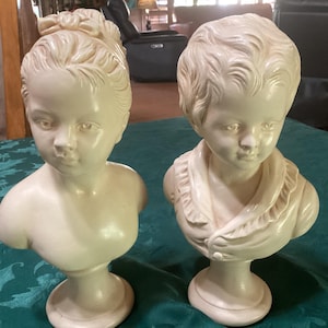 May include: Two off-white busts of children. The bust on the left has a bow in its hair. The bust on the right has short hair and a ruffled collar. Both busts are on pedestals and are likely decorative items.