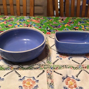 May include: Two blue ceramic bowls, one round and one rectangular, on a white tablecloth with a green floral pattern.