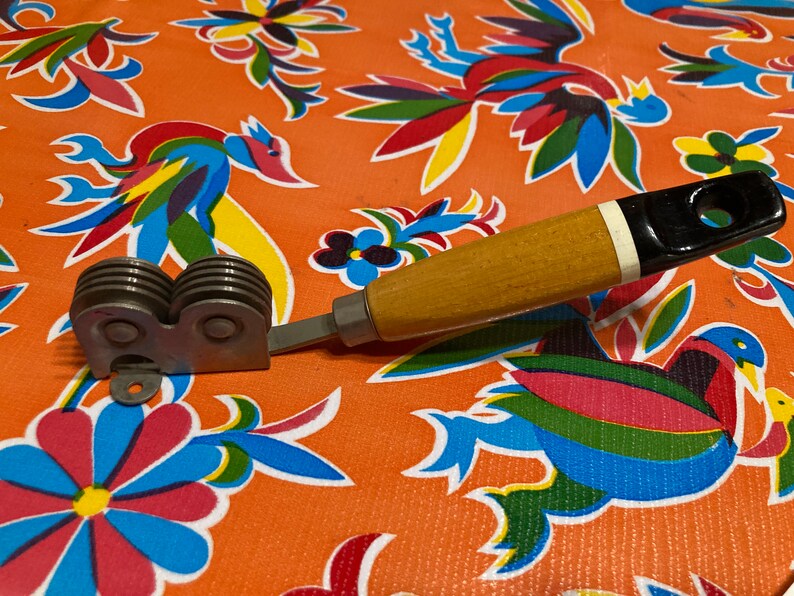 May include: A vintage kitchen tool with a wooden handle and a metal blade with multiple wheels. The handle is brown with a black and white stripe. The tool is on a colorful floral fabric.