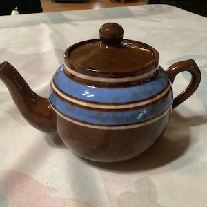 Può includere: Teiera in ceramica marrone con corpo e coperchio, con strisce orizzontali blu e bianche. La teiera ha un beccuccio curvo e un manico arrotondato. Il coperchio ha un piccolo pomello rotondo. Per preparare il tè.