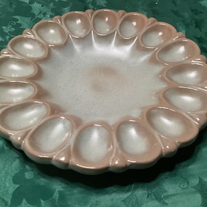 May include: A round ceramic egg serving platter featuring a smooth, glossy finish with a gradient color scheme transitioning from a soft, muted pink at the outer rim to a creamy, off-white center. The plate has eighteen molded egg-shaped indentations.