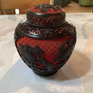 Pode incluir: Um pote de madeira esculpido vermelho e preto com uma tampa. O pote apresenta desenhos florais intrincados e um padrão geométrico.