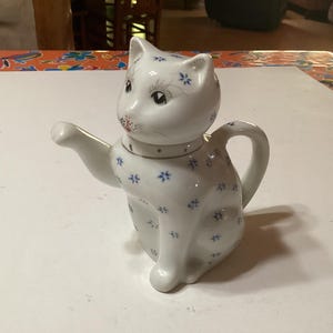 Puede incluir: Una tetera de cerámica blanca con forma de gato con diseños florales azules. La tetera tiene un pico y una manija.