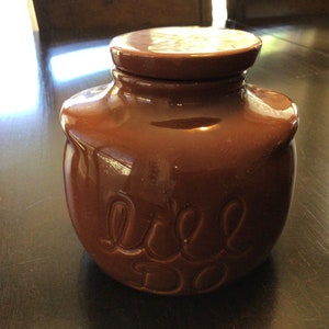 Op de afbeelding: Een bruine keramische pot met een deksel. De pot heeft de woorden "Lil' Do" in reliëf aan de voorkant.