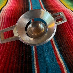 May include: A silver-colored metal object with a circular center and rectangular extensions on either side. The object has a small, spherical indentation in the center. The background is a colorful striped textile.