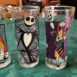 Puede incluir: Tres vasos transparentes con personajes de Pesadilla antes de Navidad. Un vaso muestra a Sally, otro a Jack Skellington en un marco de ata&uacute;d, y el tercero representa a Jack y Sally juntos con detalles de purpurina de color morado.
