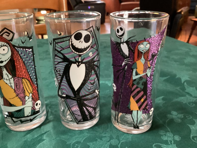 Puede incluir: Tres vasos transparentes con personajes de Pesadilla antes de Navidad. Un vaso muestra a Sally, otro a Jack Skellington en un marco de ata&uacute;d, y el tercero representa a Jack y Sally juntos con detalles de purpurina de color morado.