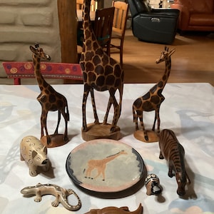 Könnte beinhalten: Eine Sammlung von Holzfiguren von Tieren, darunter Giraffen, ein Zebra, ein Krokodil und ein Flusspferd. Ein dekorativer Teller mit einer Giraffenillustration ist ebenfalls vorhanden. Die größte Giraffe ist etwa 46 cm hoch.