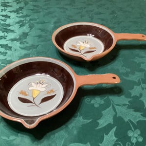 May include: Two ceramic skillets with terracotta-colored handles and rims. The interior features a dark brown border surrounding a light gray center, decorated with a white and yellow flower illustration and brown leaves, including a small pour spout.