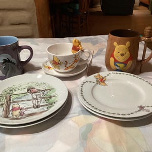 Puede incluir: Un juego de tazas de té y platillos con el tema de Winnie the Pooh, que incluye una taza marrón con un personaje de Winnie the Pooh, una taza de té blanca con un personaje de Winnie the Pooh y dos platillos blancos con personajes de Winnie the Pooh. Las tazas de té y los platillos están sobre un mantel blanco.