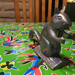 May include: A silver squirrel nutcracker with a detailed design. The nutcracker is sitting on a green tablecloth with a floral pattern.