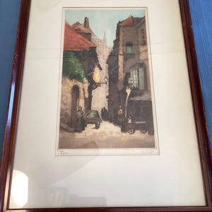 May include: A framed print of a street scene in a European city. The print is in a brown wooden frame and features a narrow street with buildings on either side. The buildings are painted in shades of brown, gray, and white. There are people walking in the street, and a horse-drawn carriage is parked in the foreground. The print is signed "Theophile Steinlen" in the lower right corner.