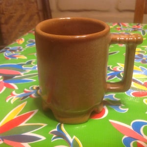 Pode incluir: Uma caneca de cerâmica marrom com uma alça. A caneca tem um fundo arredondado e uma parte superior ligeiramente alargada. Está sentada em uma toalha de mesa verde com um padrão floral.