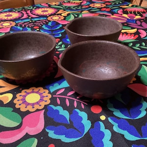 Könnte beinhalten: Drei braune Keramikschalen mit strukturierter Oberfläche stehen auf einer bunten Tischdecke mit Blumenmuster. Die Schalen haben eine einfache, abgerundete Form und einen dunklen, erdigen Farbton. Die Tischdecke zeigt eine lebendige Mischung aus Blumen und Blättern.
