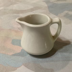 May include: A white ceramic creamer with a handle. The creamer is small and has a rounded shape.