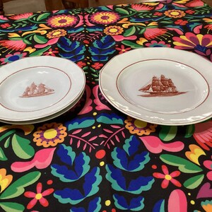 May include: Two white ceramic plates with a brown ship illustration in the center. The plates have a brown rim and are set on a colorful floral patterned tablecloth. One plate is a bowl shape, and the other is a dinner plate.