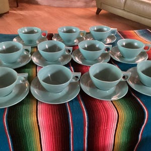 May include: A set of twelve matching aqua-colored teacups and saucers. The cups have a square handle and sit on round saucers. The set is arranged on a colorful striped textile.