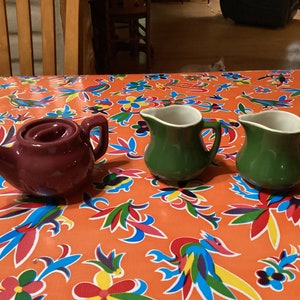 以下が含まれることがあります： カラフルなテーブルクロスの上に置かれた、セラミック製のキッチン用品3点。蓋と注ぎ口が付いた、小さな濃い紫色のティーポット。白い内側を持つ、小さな緑色のクリーマーピッチャー2つ。