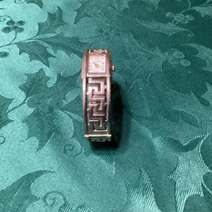 May include: Silver-toned wristwatch with a rectangular face and a Greek key design on the band. The watch face is white with black numerals and hands. The watch is displayed on a green patterned surface.