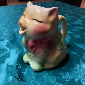 Peut inclure: Une crémière vintage en céramique en forme de chat avec un visage souriant, des joues roses et un nœud papillon rouge. Le chat est jaune clair avec des accents verts et roses. Il a une poignée et est assis droit.