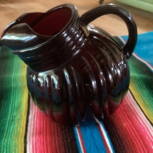 Puede incluir: Una jarra burdeos oscuro con un diseño acanalado y un asa curva. La jarra tiene un pico para verter y se encuentra sobre un textil a rayas colorido. La jarra es probablemente de cerámica o vidrio.