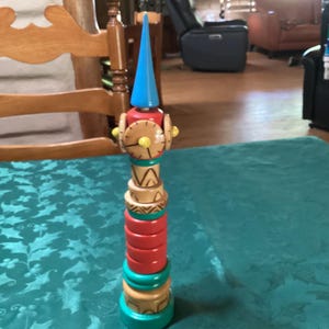 May include: A colorful wooden stacking toy with a blue cone top, red, green, and tan rings, and decorative elements. The toy is standing on a teal tablecloth. The toy is approximately 12 inches tall.