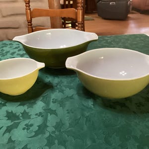 May include: A set of three glass mixing bowls with side handles. One small yellow bowl, one medium light green bowl, and one large dark olive green bowl. Each bowl features a smooth, glossy exterior finish and a clean white interior surface.