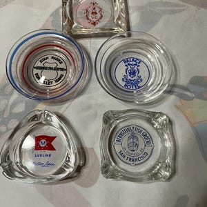 May include: A collection of five clear glass ashtrays with various designs. One square ashtray features a red and white design. Other ashtrays display text and logos, including "Utah Parks," "Palace Hotel," and "Bernstein's Fish Grotto."