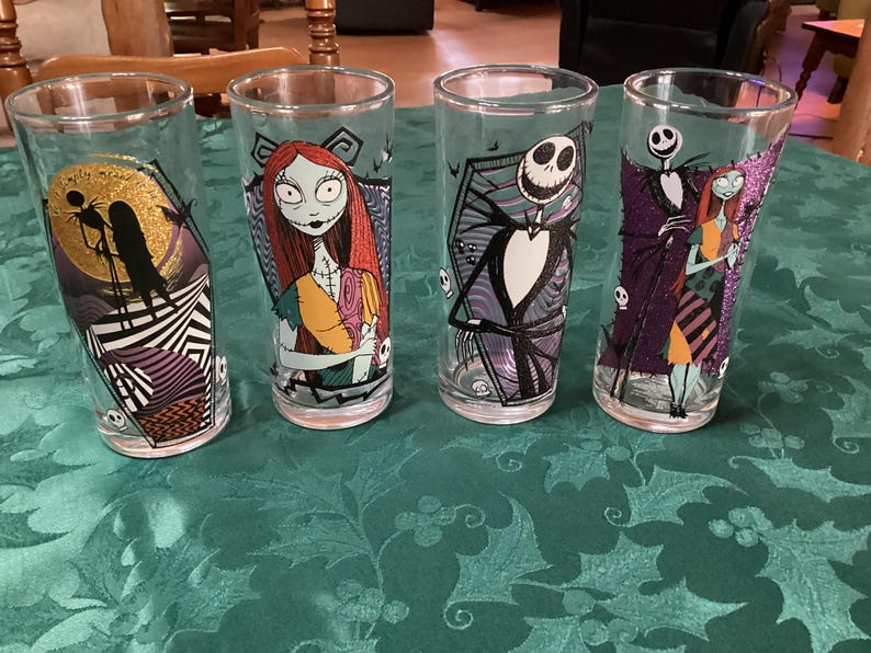 Puede incluir: Cuatro vasos de cristal transparente con personajes y escenas de Pesadilla antes de Navidad. Cada vaso muestra coloridos estampados gr&aacute;ficos de Jack Skellington y Sally, incluyendo una luna dorada con purpurina y motivos de calaveras.
