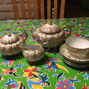 Puede incluir: Un juego de té blanco con detalles dorados. El juego incluye una tetera, una jarra de crema, un azucarero y una taza de té y un platillo. El juego de té está sobre un mantel verde con un patrón floral.