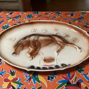 May include: A white ceramic serving platter with a brown bull design. The bull is in a running pose and has a raised tail. The platter has a scalloped edge and is decorated with black and brown accents.