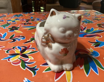 May include: White ceramic planter shaped like a cat with a gold bow and floral details. The planter is sitting on a colorful tablecloth.