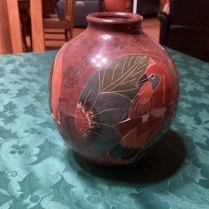 May include: A round, reddish-brown ceramic vase with a bird and leaf design. The bird is depicted in red, blue, and green, with the leaves in shades of green and blue. The vase has a glossy finish.