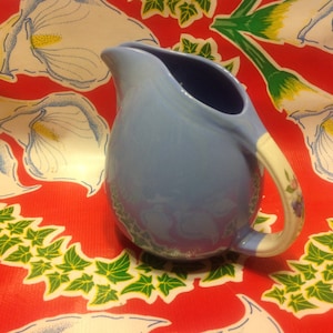 Peut inclure: Un pichet en céramique bleu clair avec une poignée blanche et un motif floral. Le pichet est posé sur une nappe rouge à motif floral blanc.