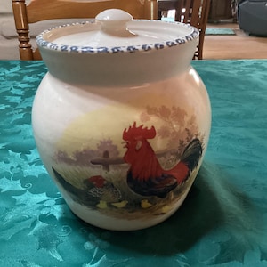 May include: A ceramic cookie jar with a lid, featuring a rooster and hen design. The jar is off-white with a blue patterned rim and a painted scene of a rooster, hen, and fence. The jar is a decorative kitchen item.